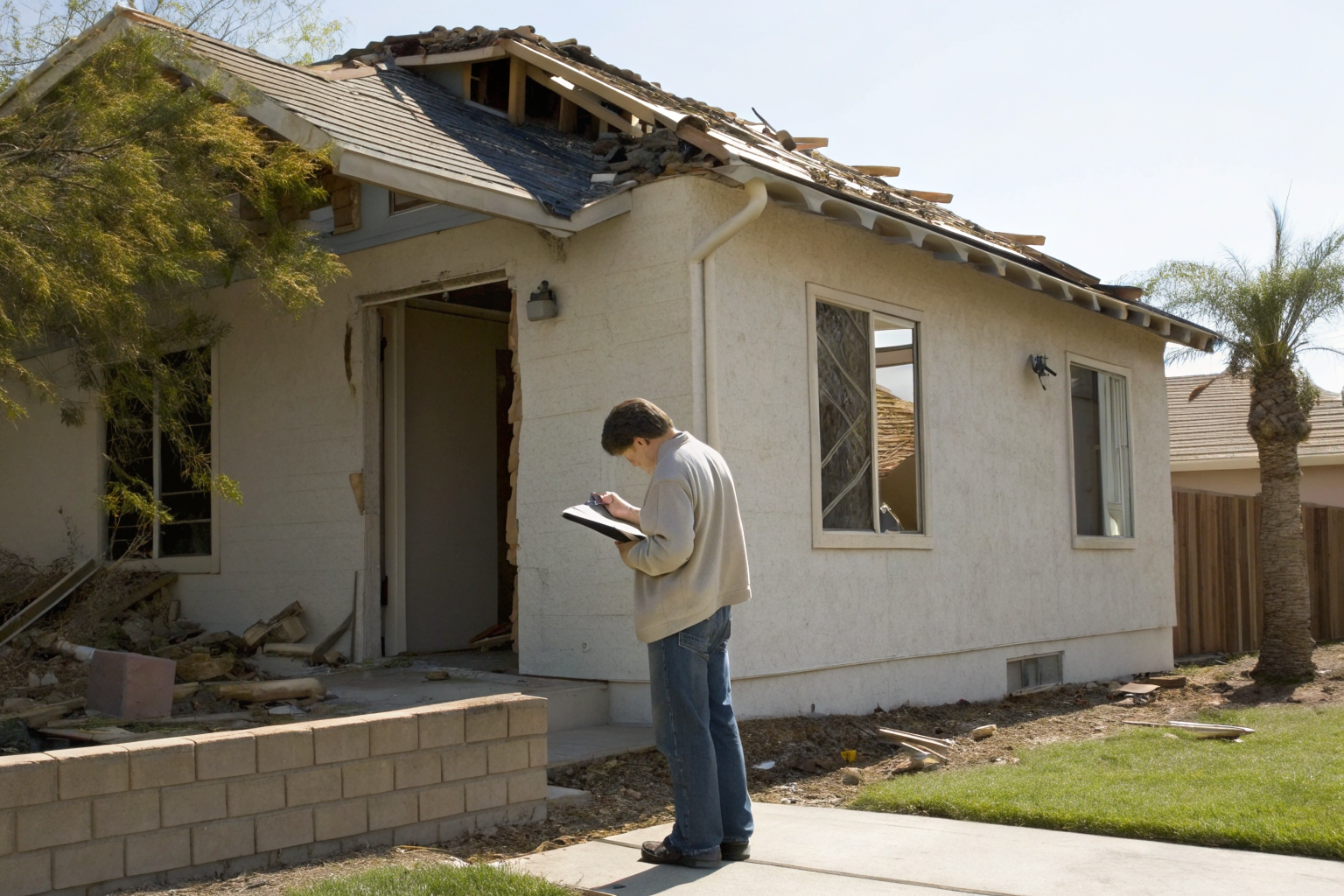 Homeowner examines storm damage on house, taking notes in daylight.