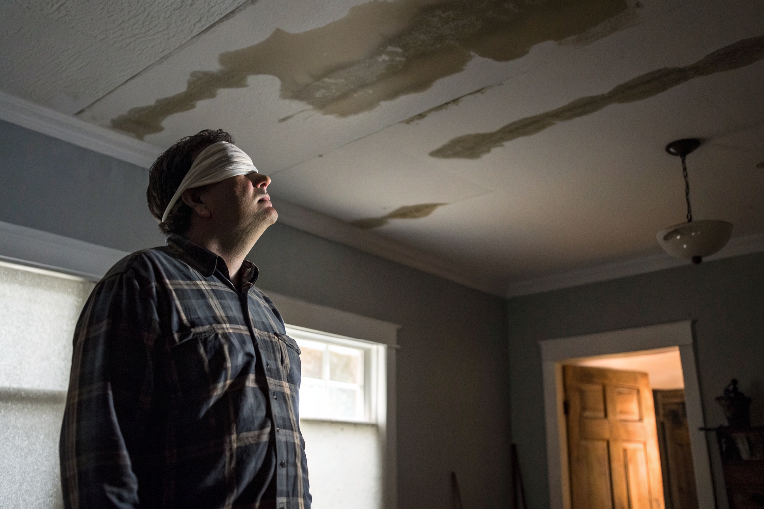 Blindfolded homeowner in a room with hidden leaks, illustrating insurance exclusions and gradual damage.